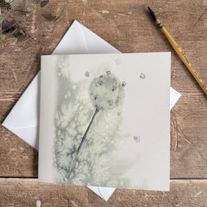 May include: Square greeting card featuring a watercolour dandelion design and paw prints. The card is white with a light green and grey illustration. An envelope is behind the card, and a paintbrush is in the background.