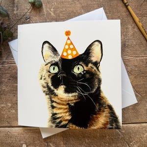 May include: A square greeting card featuring a watercolor painting of a tortoiseshell cat wearing a party hat with orange polka dots. The cat has green eyes and a black and brown coat. The card is on a white envelope.