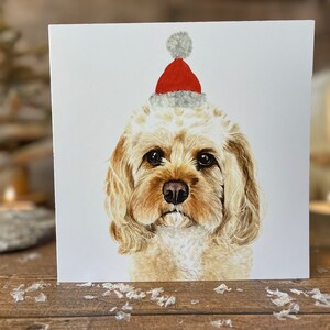 May include: A square Christmas card with a watercolour illustration of a dog wearing a red and grey Santa hat. The dog has light brown fur and a dark nose. The card is set against a white background, perfect for the festive season.