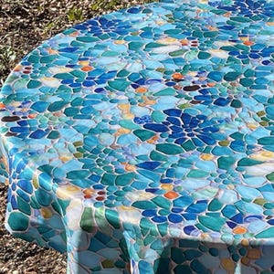 May include: Round blue and green tablecloth with a mosaic pattern. The tablecloth is draped over a round table with a white metal base.