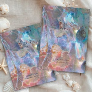 Puede incluir: Dos libros de votos de boda con cubiertas iridiscentes, con las palabras "Their Vows" y "Our Vows" en escritura elegante. Los libros están personalizados con nombres y fechas. Conchas marinas y una estrella de mar están dispuestas alrededor.