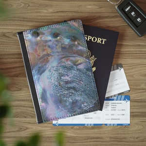 May include: A passport holder with a blue, green, and purple iridescent shell pattern. The holder is closed and sitting on top of a blue passport with the word "SPORT" printed on the cover. There are two airline boarding passes visible in the image.