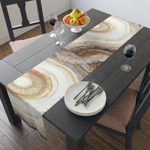 May include: A white table runner with a brown and gold abstract design. The runner is laid on a dark wood table with a white plate and silverware, a glass of wine, and a bowl of apples.