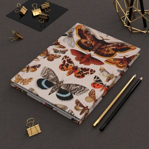 May include: A cream-colored notebook with a colorful butterfly pattern. The cover features a variety of butterflies in shades of brown, orange, red, black, and blue. The notebook is closed and lying on a gray surface.