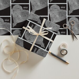 Puede incluir: Un regalo envuelto con un diseño de corazón de huellas dactilares en blanco y negro, atado con una cinta crema. El fondo presenta el mismo patrón de corazón. También hay tijeras, cinta adhesiva y un bolígrafo.