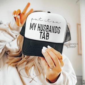 May include: A black and white trucker hat with the text "put it on my husbands tab" in black lettering. The hat has a black mesh back and a white front.