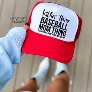 Op de afbeelding: Een rood-witte baseballcap met de tekst "Killin' this BASEBALL MOM THING" en "JUST KIDDING. WHAT INNING ARE WE IN?". De hoed wordt door een persoon omhoog gehouden.
