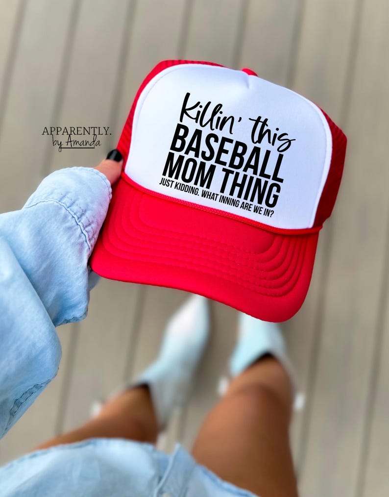 Op de afbeelding: Een rood-witte baseballcap met de tekst "Killin' this BASEBALL MOM THING" en "JUST KIDDING. WHAT INNING ARE WE IN?". De hoed wordt door een persoon omhoog gehouden.