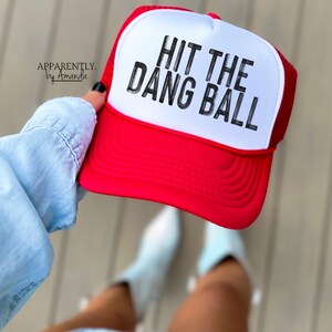 May include: Red and white trucker hat with a white mesh back. The hat has black text that reads "Hit the Dang Ball".
