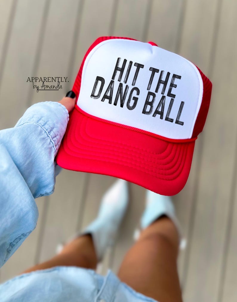 May include: Red and white trucker hat with a white mesh back. The hat has black text that reads "Hit the Dang Ball".