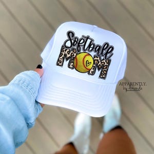 May include: White trucker hat with the words "Softball Mom" in black script and leopard print. A yellow softball with a heart is in the center. The text "Apparently. by Amanda" is on the right side of the hat.