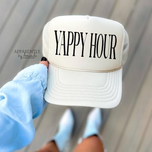 May include: Cream-colored trucker hat with the text "YAPPY HOUR" in black. The hat has a mesh back and a curved brim. The text "Apparently by Amanda" is printed on the side of the hat.