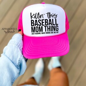 Op de afbeelding: Een felroze en witte baseballcap met de zwarte tekst "Killin' this BASEBALL MOM THING". De tekst "JUST KIDDING. WHAT INNING ARE WE IN?" staat eronder. De pet wordt door een persoon vastgehouden.