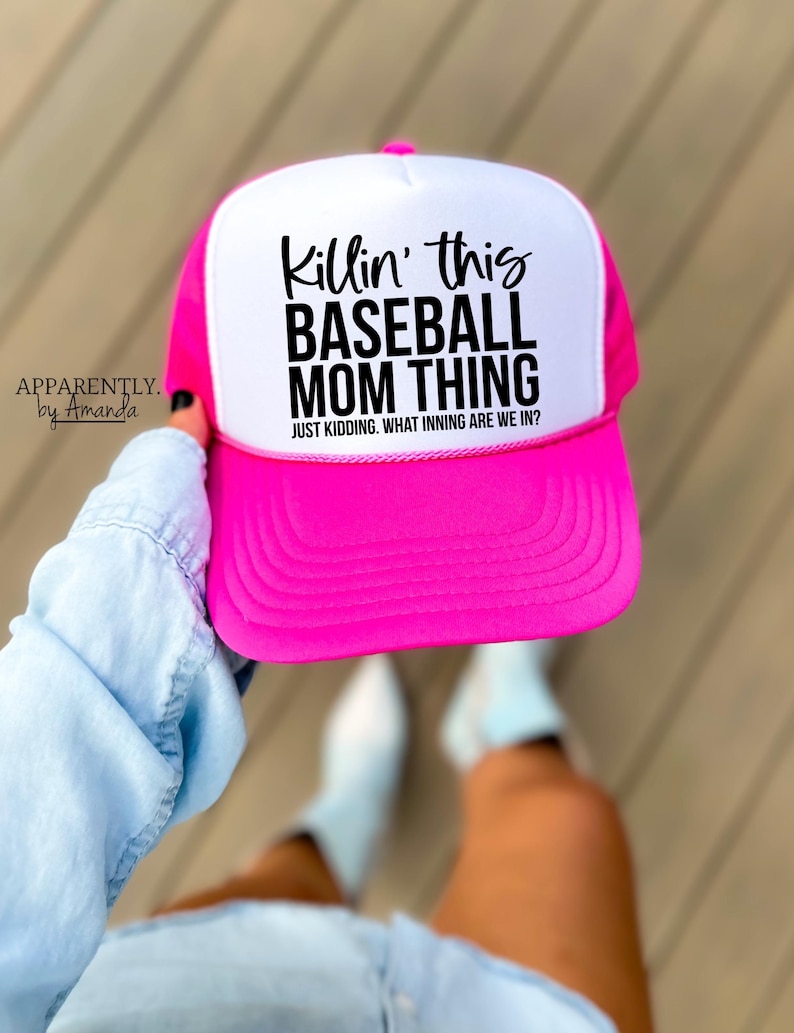 Op de afbeelding: Een felroze en witte baseballcap met de zwarte tekst "Killin' this BASEBALL MOM THING". De tekst "JUST KIDDING. WHAT INNING ARE WE IN?" staat eronder. De pet wordt door een persoon vastgehouden.