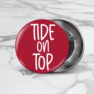 May include: A red button with the words "TIDE ON TOP" in white, handwritten-style font. The button has a shiny finish and a metal backing. The text "Apparently by Amanda" is printed at the bottom.
