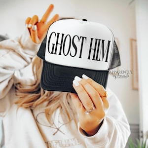 May include: A white and black trucker hat with the text "Ghost Him" in black lettering. The hat has a black mesh back and a white front panel.