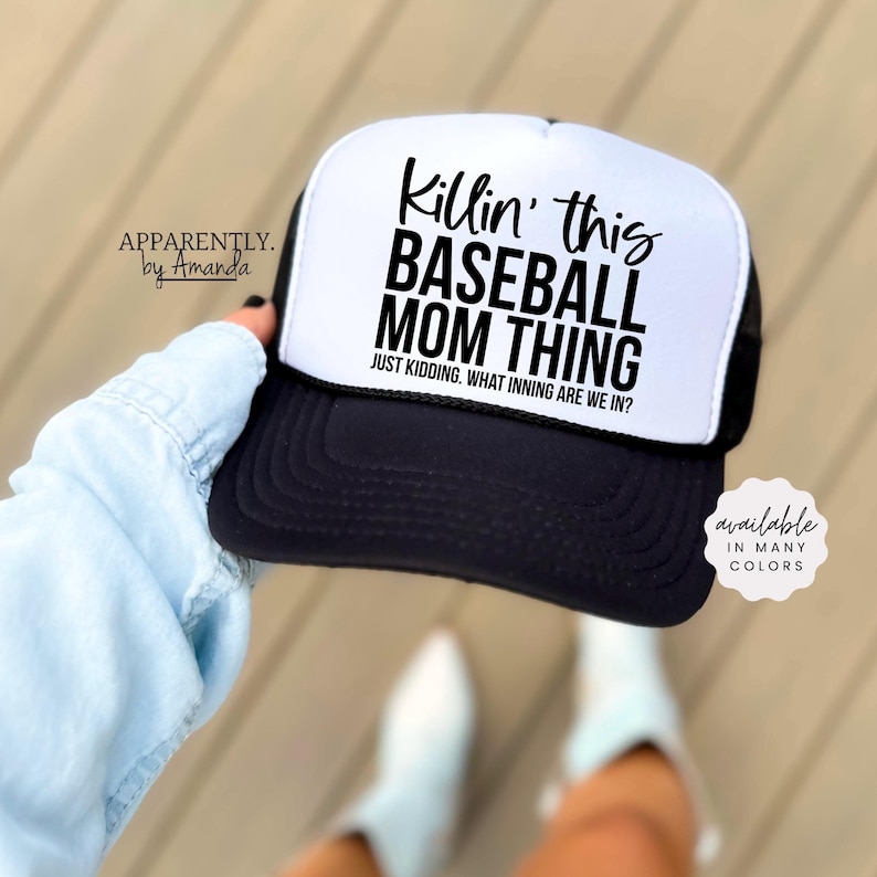 Op de afbeelding: Zwart-witte baseballcap met de tekst "Killin' this BASEBALL MOM THING" en "JUST KIDDING. WHAT INNING ARE WE IN?". De hoed is een trucker-stijl met een mesh rug en een gebogen klep. De tekst "available in many colors" is ook zichtbaar.