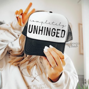 May include: A black and white trucker hat with the text "completely unhinged" in black script. The hat has a black mesh back and a white front panel. The brand name "APPARENTLY. by Amanda" is printed in black on the back of the hat.