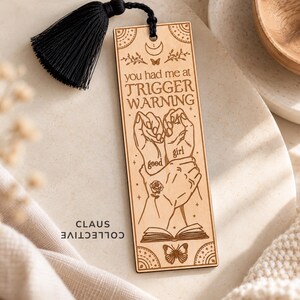 May include: A rectangular light-colored wooden bookmark featuring a black tassel at the top. The design includes engraved illustrations of hands, a rose, a butterfly, and an open book, with the text you had me at TRIGGER WARNING good girl.