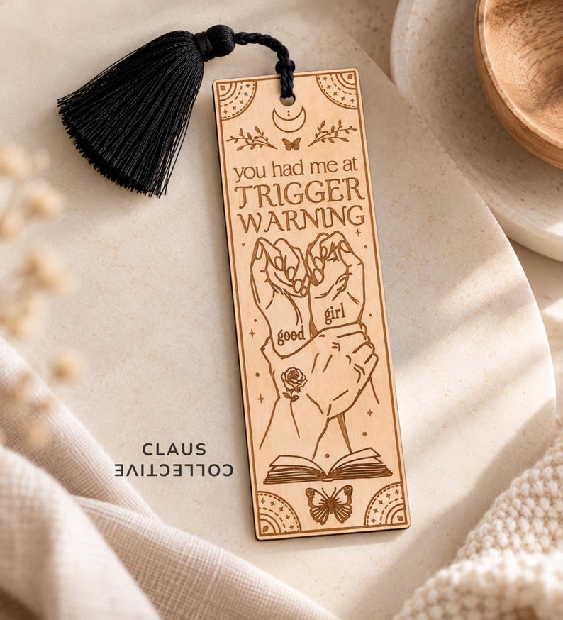 May include: A rectangular light-colored wooden bookmark featuring a black tassel at the top. The design includes engraved illustrations of hands, a rose, a butterfly, and an open book, with the text you had me at TRIGGER WARNING good girl.