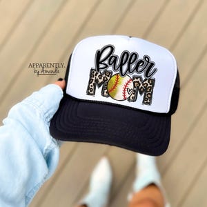 May include: A black and white trucker hat with the words "Baller Mom" in a script font and leopard print accents. A baseball with a heart is in the center. The hat has a black brim and mesh back. The text "Apparently. by Amanda" is on the side.
