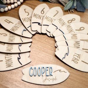 May include: Wooden surfboard-shaped monthly milestone markers with the numbers 1 through 12 and the word "month" or "year" printed on each. The markers are arranged in a circle around a smaller surfboard-shaped sign with the name "COOPER Joseph" printed on it.