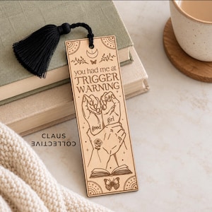 May include: A rectangular wooden bookmark featuring a black tassel and laser-engraved illustrations of hands, a rose, a butterfly, and an open book. The text reads: you had me at TRIGGER WARNING, with the words good girl written on the hands.