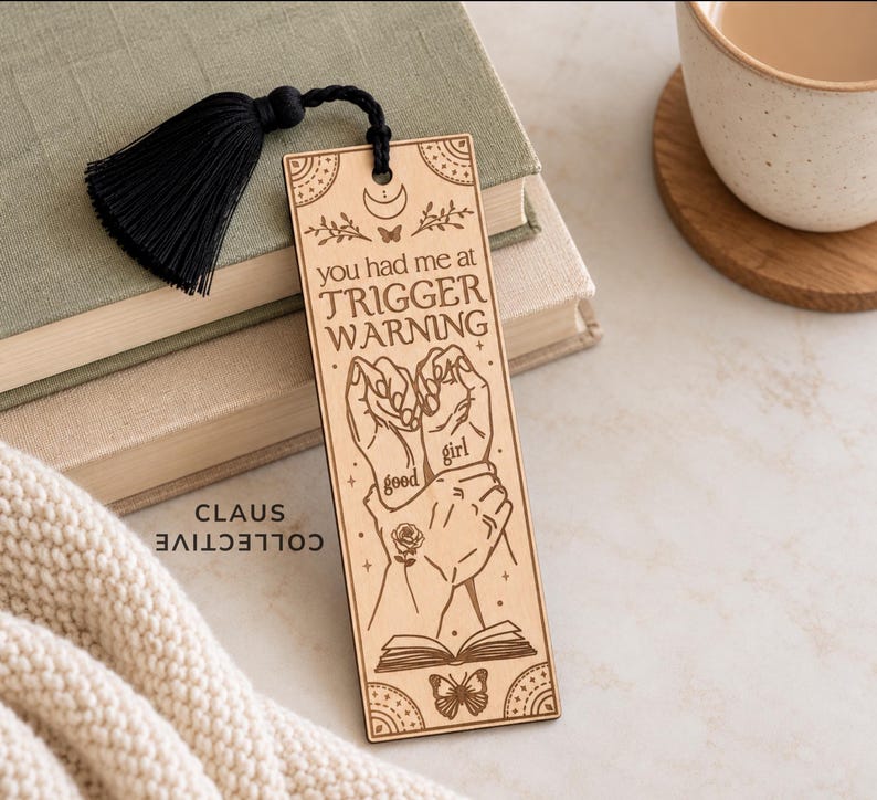 May include: A rectangular wooden bookmark featuring a black tassel and laser-engraved illustrations of hands, a rose, a butterfly, and an open book. The text reads: you had me at TRIGGER WARNING, with the words good girl written on the hands.