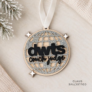 May include: A circular wooden ornament featuring a silver glitter globe design with four small white stars. The black text reads dwts couch judge. A white ribbon is attached to the top for hanging. The ornament has a natural wood border.