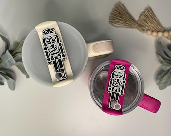 Può includere: Due bicchieri isolanti con coperchi, uno color crema e uno magenta. Ogni coperchio presenta un disegno di schiaccianoci in bianco e nero. I bicchieri hanno manici colorati abbinati su sfondo bianco.