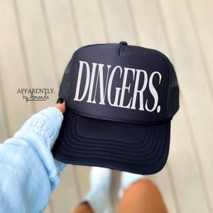 May include: A black trucker hat with a white mesh back. The hat has the word "DINGERS" printed in white on the front.