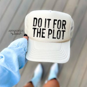 May include: Cream-colored trucker hat with the text "DO IT FOR THE PLOT" in black. The hat has a mesh back and a curved brim. The brand name "Apparently. by Amanda" is printed on the side.