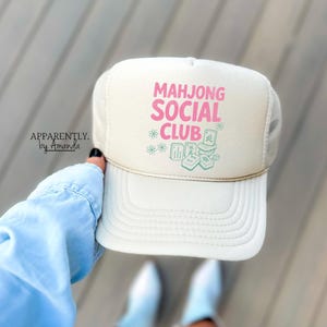 May include: A cream-colored trucker hat featuring the pink text "MAHJONG SOCIAL CLUB" above a graphic of green mahjong tiles and small flower icons. The hat has a structured front panel, a curved brim, and a decorative gold cord across the base.