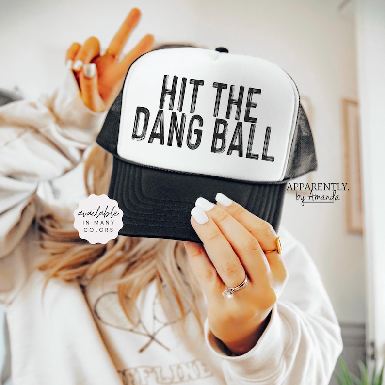 May include: A black and white trucker hat with the text "HIT THE DANG BALL" in black lettering. The hat is available in many colors.