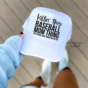 Op de afbeelding: Witte baseballcap met een mesh achterkant en een gebogen klep. De cap heeft de tekst "Killin' this BASEBALL MOM THING" in zwart, met de zin "JUST KIDDING. WHAT INNING ARE WE IN?". De merknaam "APPARENTLY. by Amanda" is ook zichtbaar.