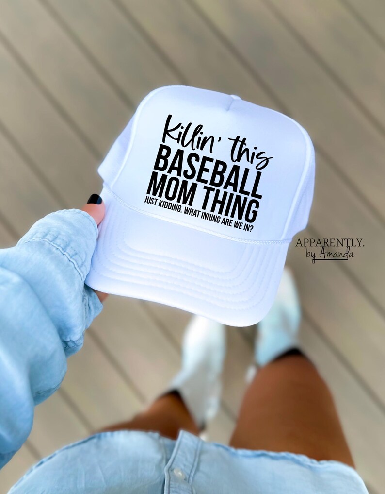 Op de afbeelding: Witte baseballcap met een mesh achterkant en een gebogen klep. De cap heeft de tekst "Killin' this BASEBALL MOM THING" in zwart, met de zin "JUST KIDDING. WHAT INNING ARE WE IN?". De merknaam "APPARENTLY. by Amanda" is ook zichtbaar.