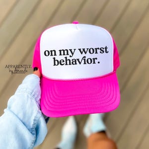 May include: A pink and white trucker hat with the text "on my worst behavior." printed in black.
