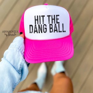 May include: A pink and white trucker hat with the text "HIT THE DANG BALL" in black lettering.