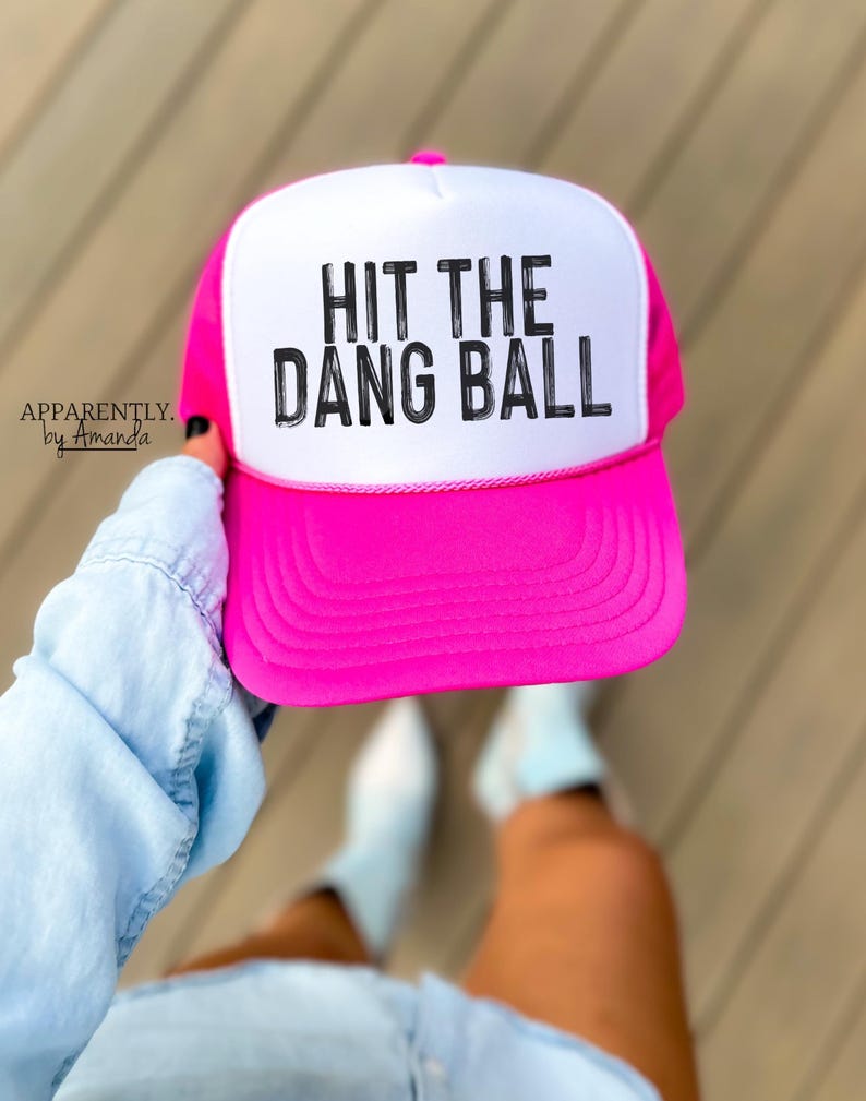 May include: A pink and white trucker hat with the text "HIT THE DANG BALL" in black lettering.
