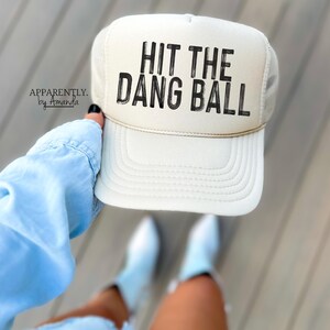 May include: A white trucker hat with a black mesh back. The hat has black text that reads "HIT THE DANG BALL".
