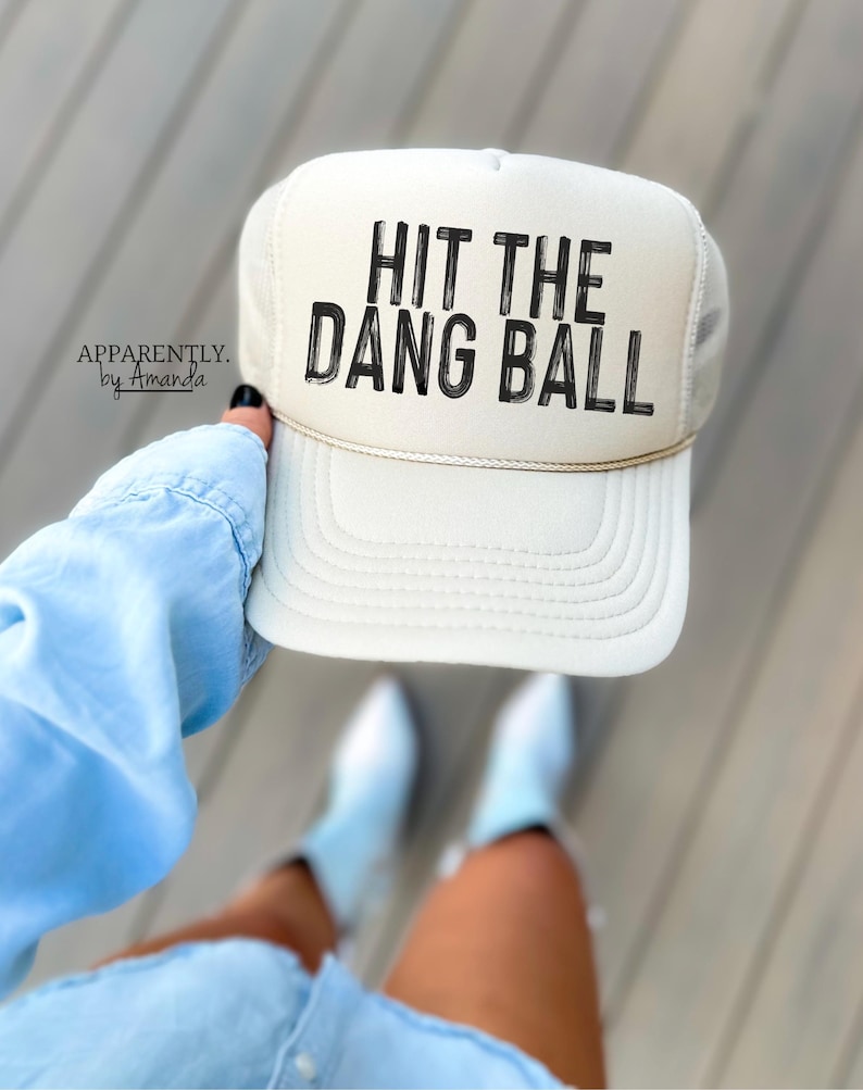 May include: A white trucker hat with a black mesh back. The hat has black text that reads "HIT THE DANG BALL".