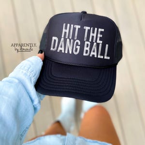 May include: Black trucker hat with a white mesh back. The hat has white text that reads "Hit the Dang Ball".