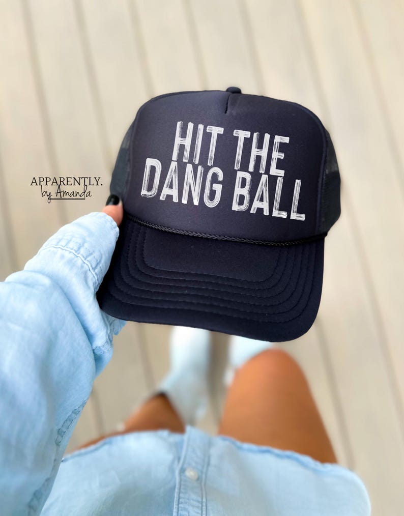 May include: Black trucker hat with a white mesh back. The hat has white text that reads "Hit the Dang Ball".