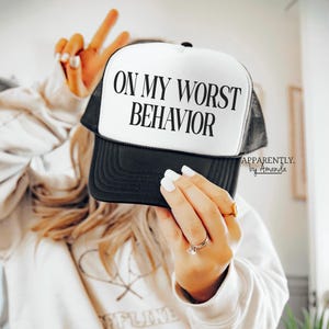 May include: A white and black trucker hat with the text "On My Worst Behavior" in black lettering. The hat has a black mesh back and a white front.