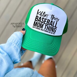 Op de afbeelding: Een groene en witte baseballcap met de zwarte tekst "Killin' this BASEBALL MOM THING". De tekst "JUST KIDDING. WHAT INNING ARE WE IN?" staat eronder. De pet wordt vastgehouden door een persoon die een lichtblauw spijkeroverhemd draagt.