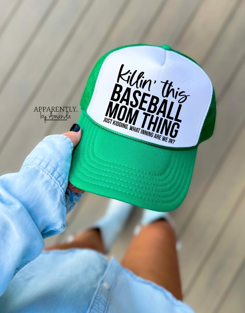 Op de afbeelding: Een groene en witte baseballcap met de zwarte tekst "Killin' this BASEBALL MOM THING". De tekst "JUST KIDDING. WHAT INNING ARE WE IN?" staat eronder. De pet wordt vastgehouden door een persoon die een lichtblauw spijkeroverhemd draagt.