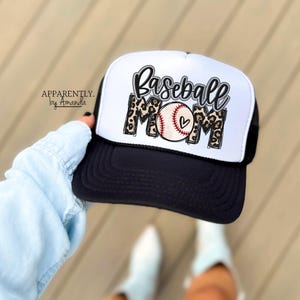 May include: A black and white baseball cap with the words "Baseball Mom" in a script font and leopard print accents. The baseball graphic has a heart. The hat has a black mesh back and a white front.