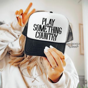 May include: A black and white trucker hat with the text "PLAY SOMETHING COUNTRY" in bold, black letters. The hat has a black brim and mesh back. The words "APPARENTLY. by Amanda" are printed in the lower right corner.