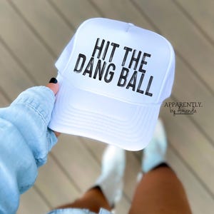 May include: White trucker hat with black text that reads "Hit the Dang Ball". The hat is held by a person wearing a light blue shirt and denim shorts.