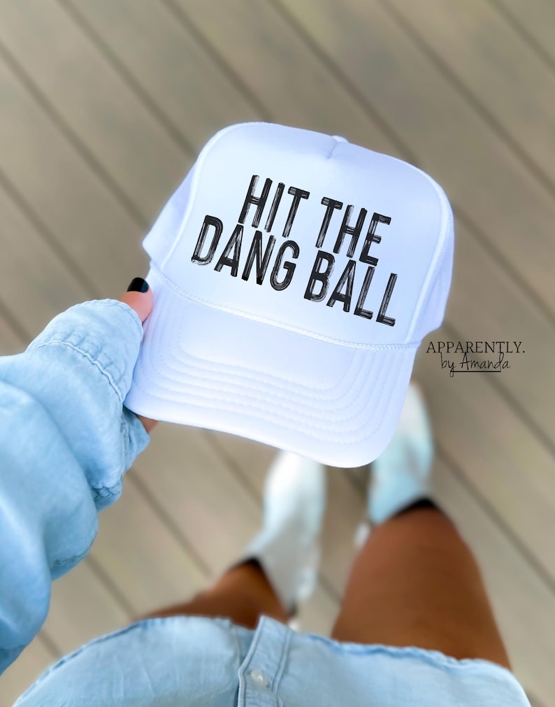 May include: White trucker hat with black text that reads "Hit the Dang Ball". The hat is held by a person wearing a light blue shirt and denim shorts.